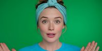 Young woman with turquoise headband and blue top gesturing with hands against bright green background, expressing surprise or explanation.
