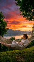 Woman relaxing in hammock during golden hour sunset with mountain lake view. Peaceful evening leisure time outdoors surrounded by lush green trees.