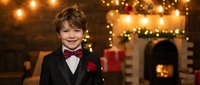 Young Caucasian boy in formal black tuxedo with red bow tie and boutonniere smiling at elegant holiday party with warm string lights and fireplace.