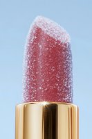 Frozen red lipstick covered in frost crystals with golden metallic case against light blue background, macro photography showing detailed texture and sparkle.