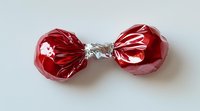 Red candy wrapped in metallic foil with twisted ends on white background, isolated sweet treat ready for design use in confectionery and holiday projects.