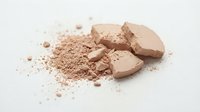 Crushed beige powder makeup with broken compact pieces scattered on white surface, suitable for cosmetics, beauty products, and skincare advertising.
