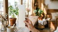 Mature man holding blank white vertical card in warm home interior with potted plants and natural sunlight for mockup and branding purposes.