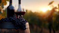 Red wine glasses and bottles with fresh grapes on wooden barrel against sunset vineyard background, romantic evening atmosphere in winery.