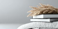 Minimalist autumn composition with dried pampas grass on stack of white books and knitted sweater against light gray background. Cozy home decor styling.