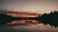 Solitary boat floating on calm lake at sunset with silhouetted forest treeline reflecting in still water, peaceful nature landscape for travel and relaxation themes.