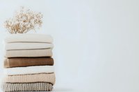 Neatly folded stack of cozy knit sweaters and cardigans in neutral beige and brown tones with dried flowers on white background, minimalist winter wardrobe styling.