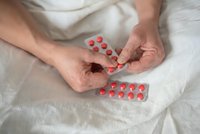 Senior hands holding red medication pills in blister pack on white bedding for healthcare and treatment concepts.