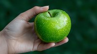 Fresh green apple with water drops held in feminine hand against dark green background, showcasing healthy organic produce for food concepts.