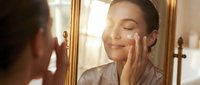 Young woman with olive skin tone and dark hair applying skincare product to her face while looking in a mirror, suitable for beauty, wellness, and cosmetics marketing.