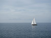 Sailboat gliding on calm blue sea under overcast sky with distant mountains on horizon, evoking freedom and adventure.