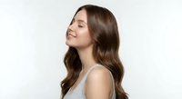 Young Caucasian woman with long wavy brown hair in gray tank top smiling peacefully with eyes closed against white background for wellness and beauty concepts.