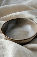 Handcrafted ceramic bowl with rustic glazed finish on soft white linen fabric, showcasing organic textures and natural earthen tones in minimalist style.