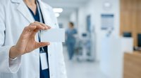 Doctor in white coat holding blank business card in modern hospital clinic setting with blurred medical staff and equipment in background.