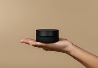 Female hand holding black round cosmetic jar against neutral beige background, minimalist product photography for beauty and skincare marketing.