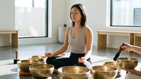 Woman of East Asian descent in meditation pose surrounded by singing bowls in bright studio space for wellness and mindfulness content.