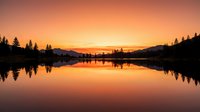 Serene lake at sunset with golden sky reflecting on calm water, surrounded by evergreen forest silhouettes and distant mountains, ideal for nature and landscape projects.