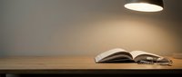 Open book with eyeglasses on wooden desk under warm pendant light, suitable for education, reading, learning, and study concepts.