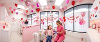 Dental office decorated for Valentine's Day with pink heart balloons, female dentist in scrubs examining young patient in festive clinical setting.