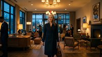Professional blonde businesswoman in elegant black coat standing confidently in upscale hotel lobby with fireplace and city views at evening.