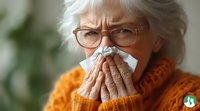Senior woman in orange sweater blowing nose with tissue, showing cold and flu symptoms against blurred green background. Concept for healthcare and illness.
