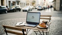 Laptop with coffee cup and croissant on wooden chair at outdoor urban street cafe, freelance work setup for remote business and lifestyle content.