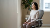 Young Asian woman in beige outfit sitting peacefully in modern chair by window, eyes closed in meditation or relaxation at home with natural light.