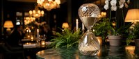 Elegant hourglass with golden metal frame sits on marble table in luxurious restaurant interior with warm ambient lighting and greenery creating sophisticated atmosphere.
