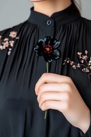 Elegant female hand holding single black cosmos flower against dark pleated blouse with delicate floral embroidery, creating dramatic gothic romantic mood.