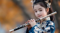 Young Asian girl in traditional blue floral dress playing ornate flute with decorative hair accessories against warm blurred background for cultural music education.