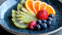 Fresh mixed fruit plate with sliced kiwi, orange segments, strawberry, raspberry and blueberries on blue ceramic plate with water droplets, macro food photography.