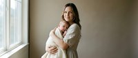 Young Caucasian mother in beige cardigan tenderly holding newborn baby by window in warm natural light creating intimate bonding moment for family lifestyle content.