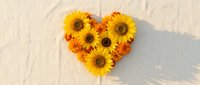 Heart-shaped arrangement of bright yellow sunflowers and orange marigolds on sandy beach surface with natural shadows for romantic summer designs.