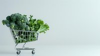 Fresh green broccoli and kale leaves in miniature shopping cart on light background, healthy eating and grocery shopping concept for social media.