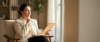 Professional Asian businesswoman reading book while sitting in comfortable chair by window in modern home office during daytime relaxation break.