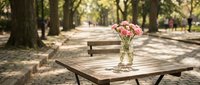 Wooden picnic table with pink flowers in glass vase in sunny park alley lined with trees, ideal for outdoor dining and leisure activities.