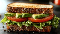 Fresh sandwich with whole grain bread, crisp lettuce, ripe tomato slices and creamy avocado on dark background, close up view of healthy lunch option.