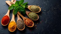Colorful spices and herbs in wooden spoons with fresh mint on dark slate background for culinary cooking and seasoning recipes.