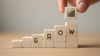 Hand placing wooden block with growth chart icon on ascending stair-like arrangement of blocks spelling "GROW" to represent business development and success.
