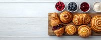 Fresh baked pastries and sweet buns on wooden board with berry toppings and cream sauce, served on white wooden table background with copy space.