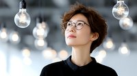 Young Asian businesswoman in glasses looking up at hanging light bulbs symbolizing creative ideas, innovation and inspiration in modern office.