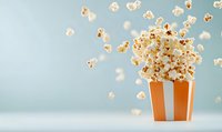 Fresh popcorn exploding from striped orange and white container against light blue background, creating dynamic motion of scattered kernels in air.
