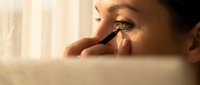 Young woman applying black eyeliner makeup in mirror with natural window light creating warm golden hour beauty routine moment.
