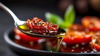 Sun-dried tomato drizzled with olive oil and seasoning on metal spoon against dark background, macro food photography with selective focus and natural lighting.