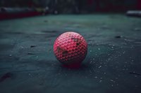 Pink golf ball on dark green surface with dramatic lighting and moody atmosphere, creating contrast between vibrant ball texture and grungy background.
