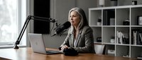 Professional middle-aged Caucasian businesswoman recording podcast in modern office with microphone and laptop for content creation and broadcasting.