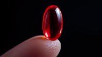 Red transparent pill capsule on fingertip against dark background, macro pharmaceutical photography showing medicine and healthcare concept.