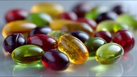Colorful vitamin and fish oil supplements scattered on reflective surface with soft focus effect, showing translucent gel capsules in red, green and amber colors.