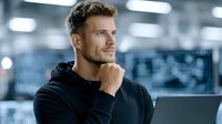 Young Caucasian businessman in black hoodie thinking while working on laptop in modern office environment with blurred background lighting.