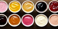 Colorful assortment of dipping sauces and condiments in white ceramic bowls arranged in two rows on dark wooden surface, top view panoramic composition.